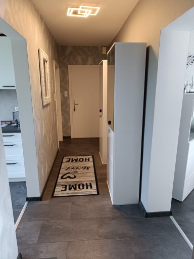 Langer Flur mit weißer Wand, grauem Boden und einem Teppich mit der Aufschrift "HOME".